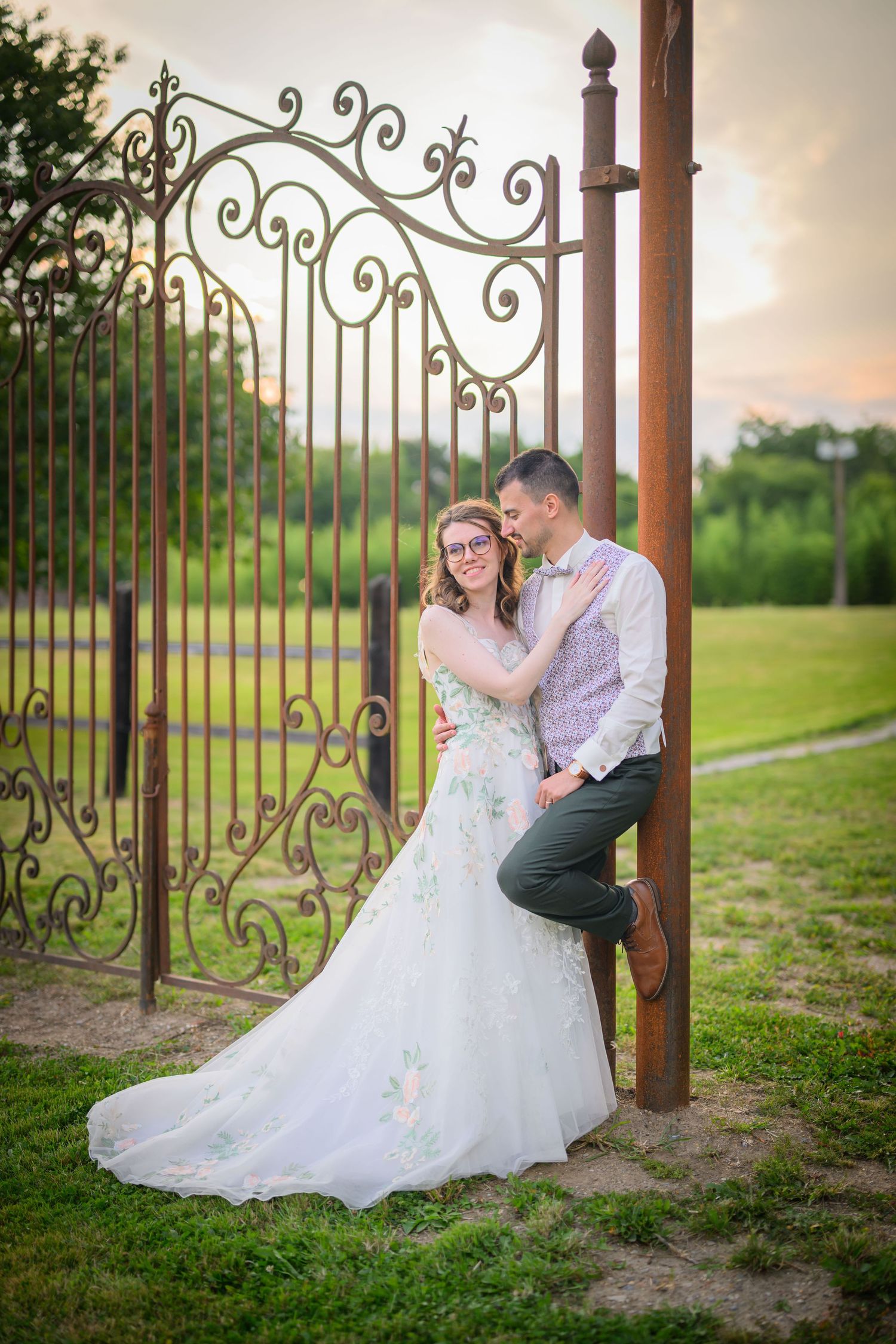 Wedding couple poses romantically by ornate iron gate during golden hour.