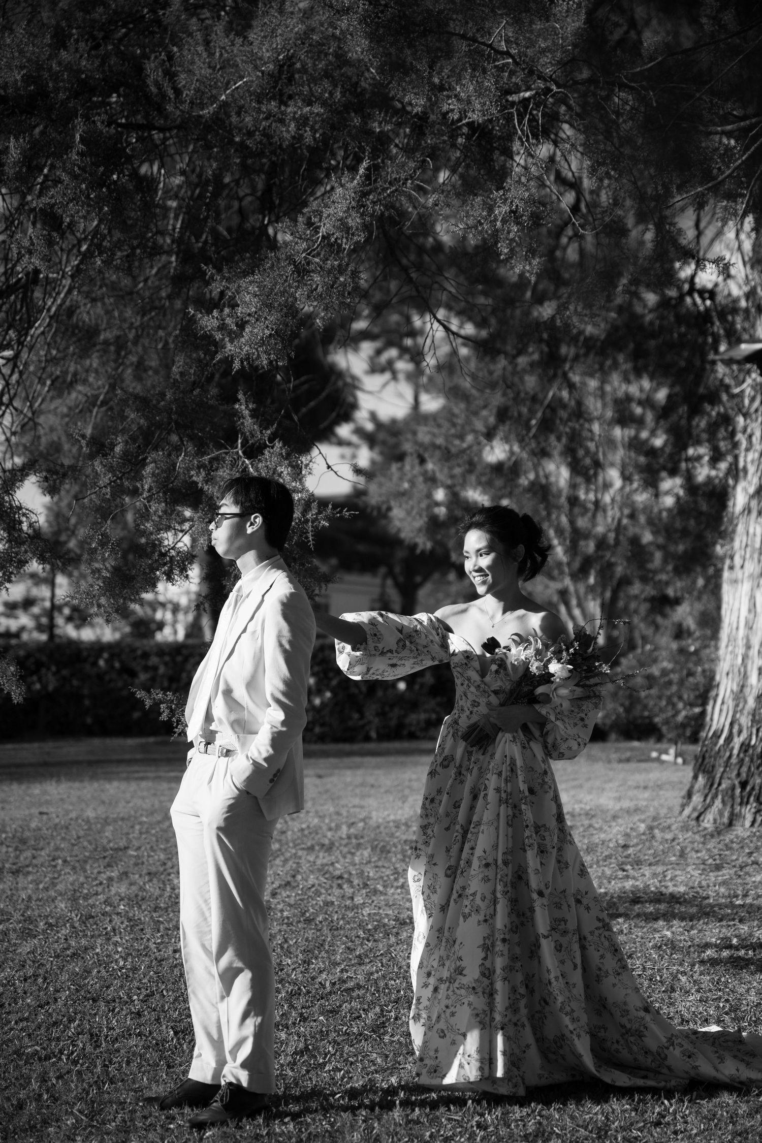 Black and white photo of bride in floral gown reaching for groom under tall pine trees.