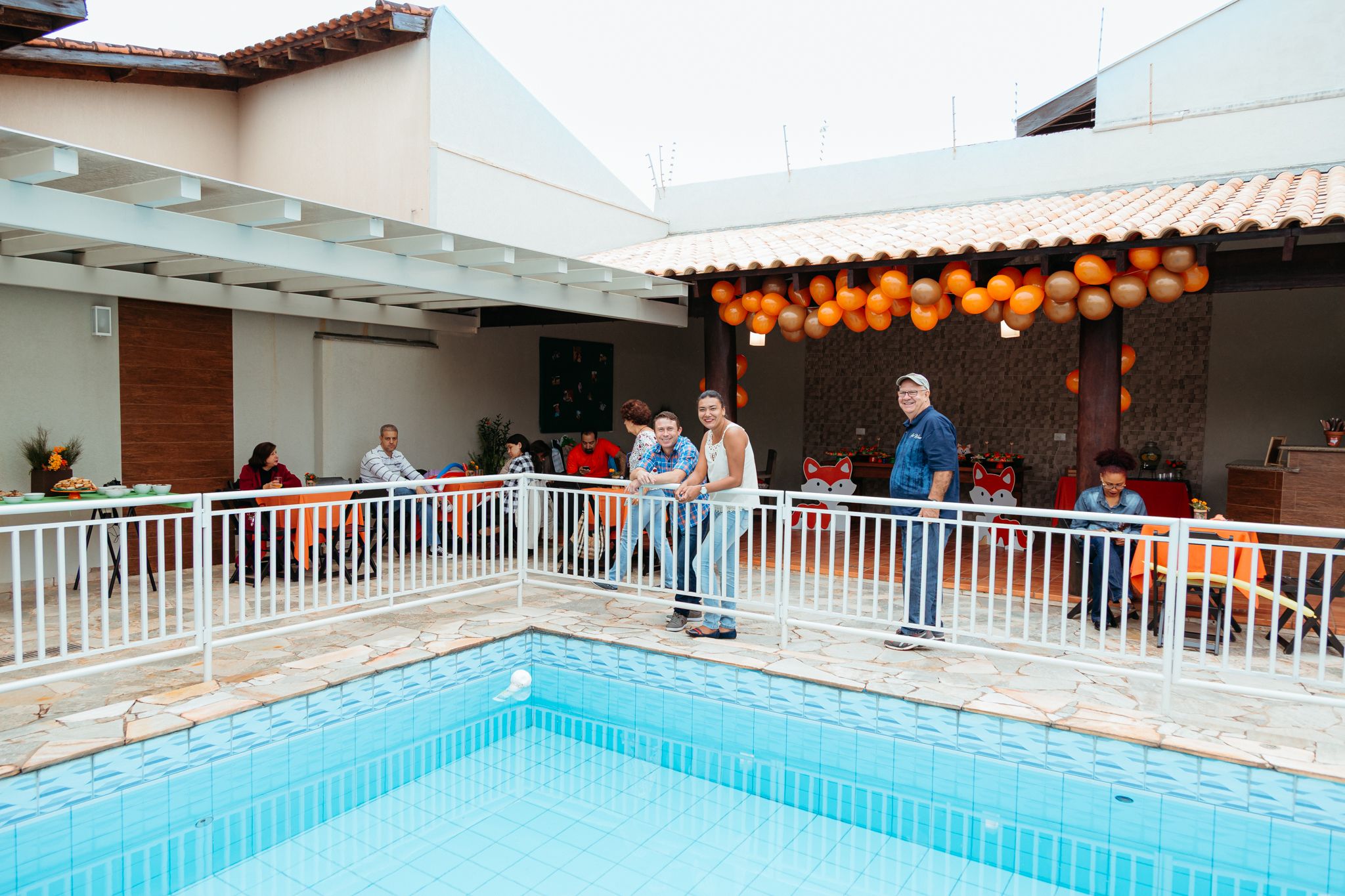 People gather around a swimming pool on a covered deck decorated with glowing orange lanterns at an evening event.