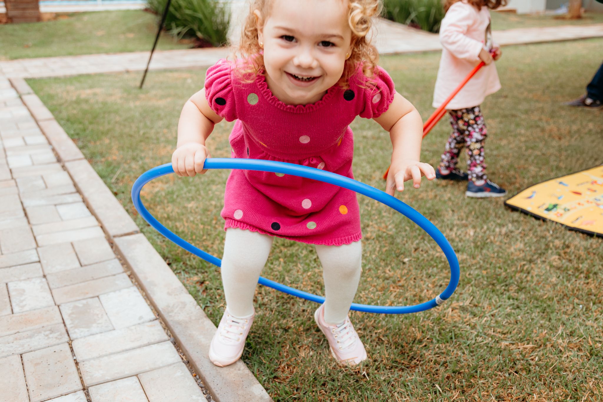 Child playing with blue hula hoop on brick pathway in yard while wearing bright pink shirt and white shoes.