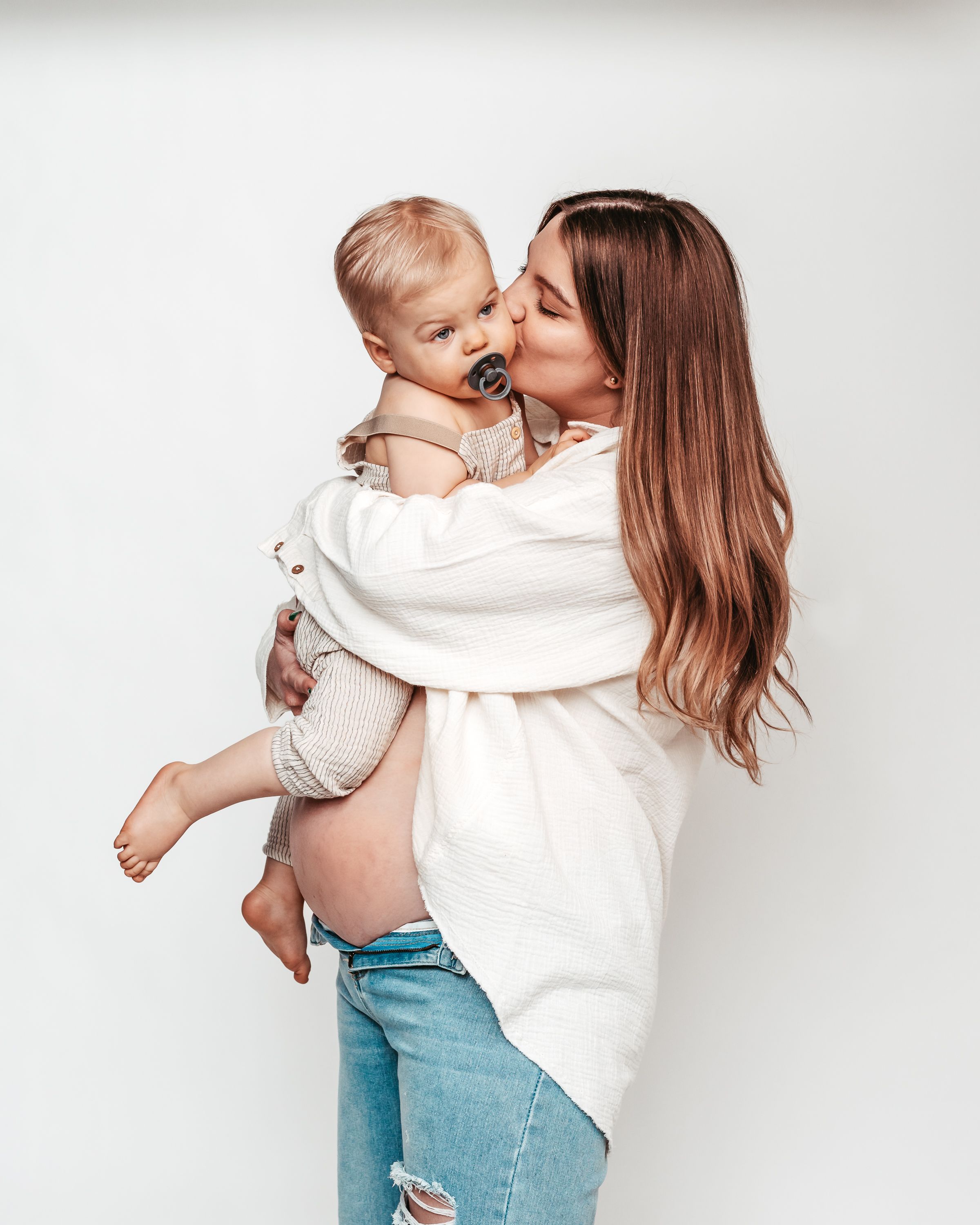 A parent in a white sweater and jeans shares tender moments with a baby against a bright white background.