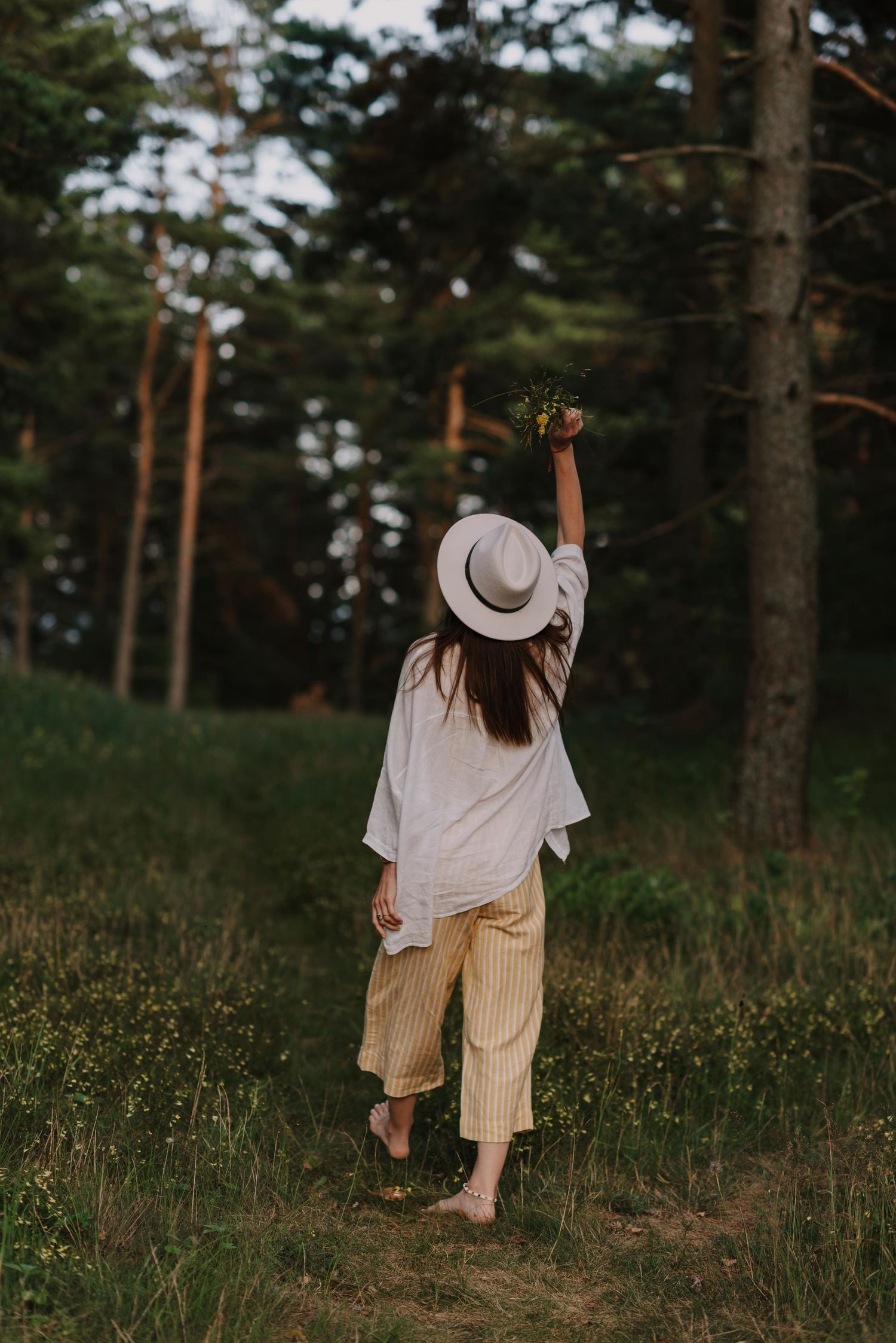 A person in a sun hat and striped pants walks through tall grass in a forest clearing.