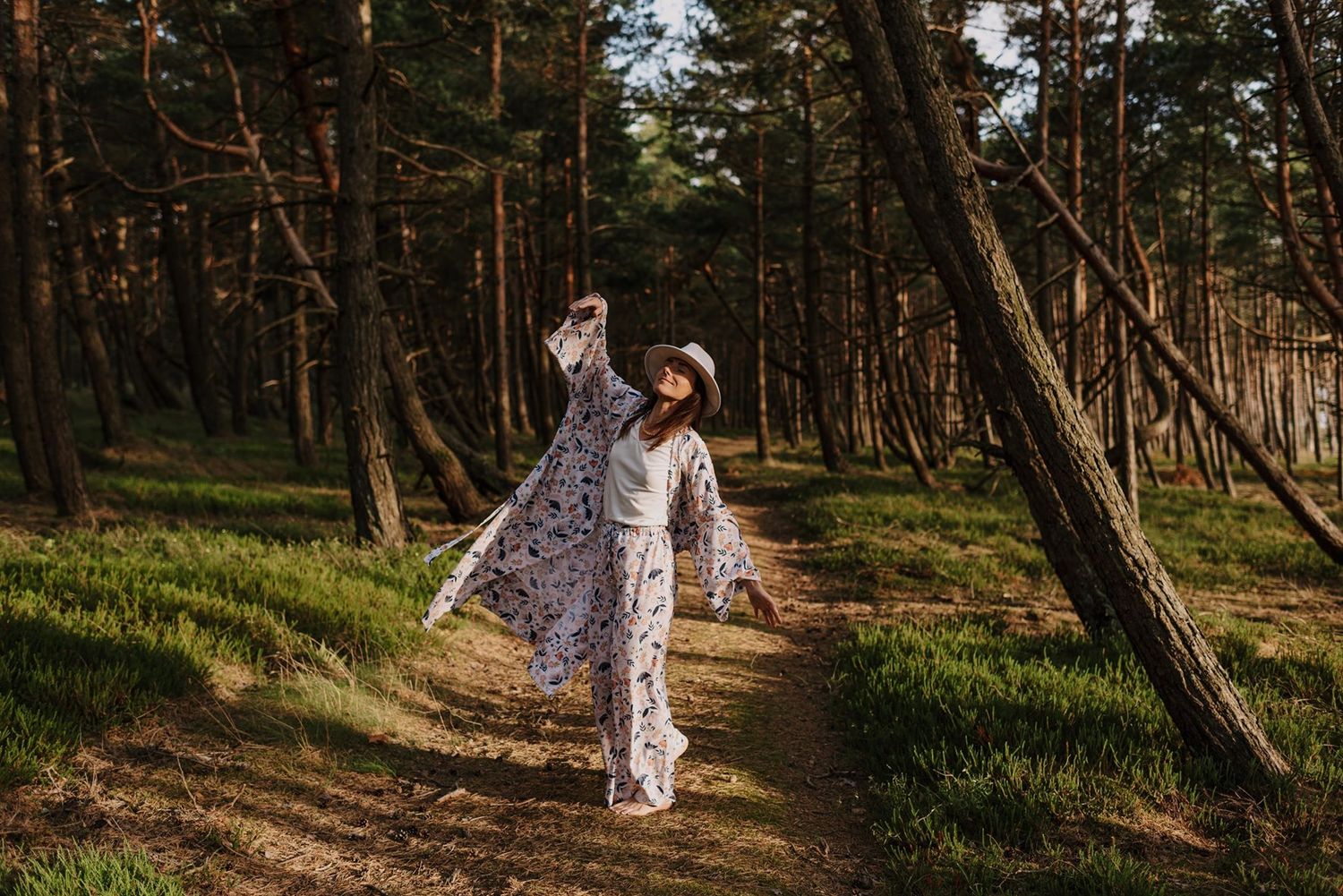 A white floral dress stands out against a winding forest path lined with pine trees.