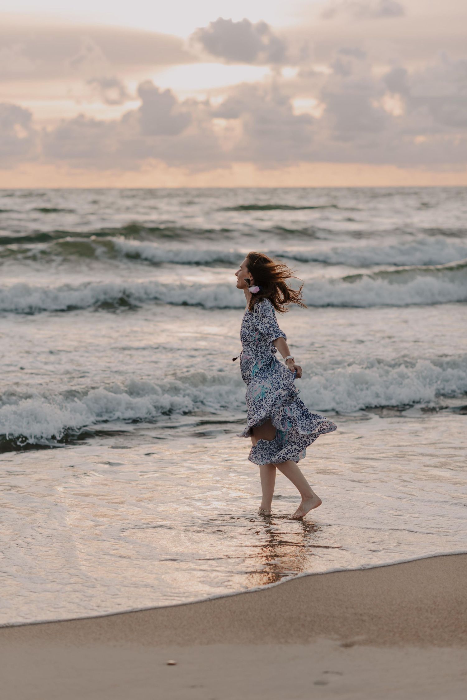 Person in a flowing dress runs playfully along a beach shoreline during sunset as waves crash nearby.