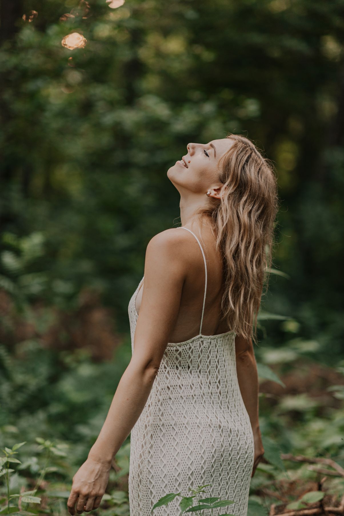 A woman in a white crochet dress looks up at sunlight filtering through forest trees.
