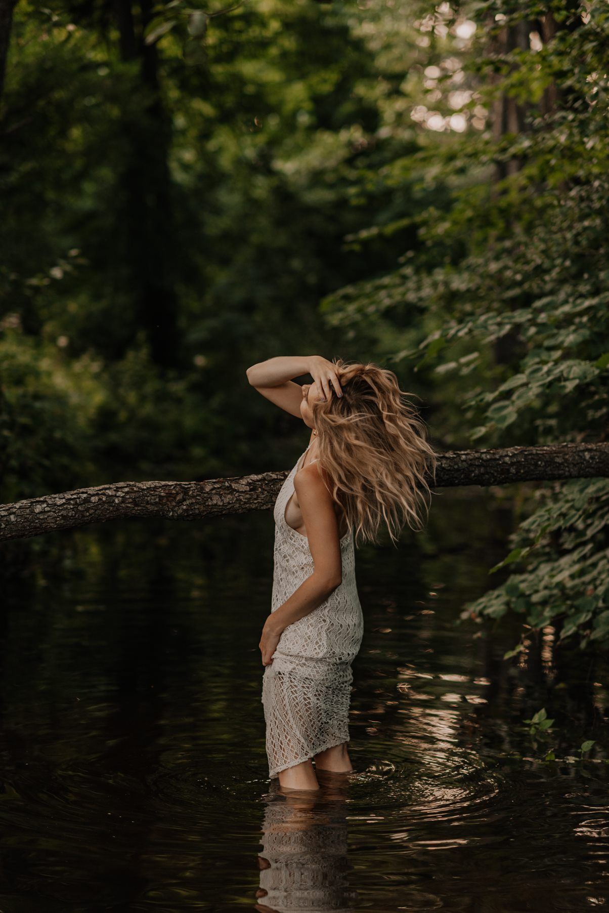 Figure in white dress wading through dark waters under tree branches.