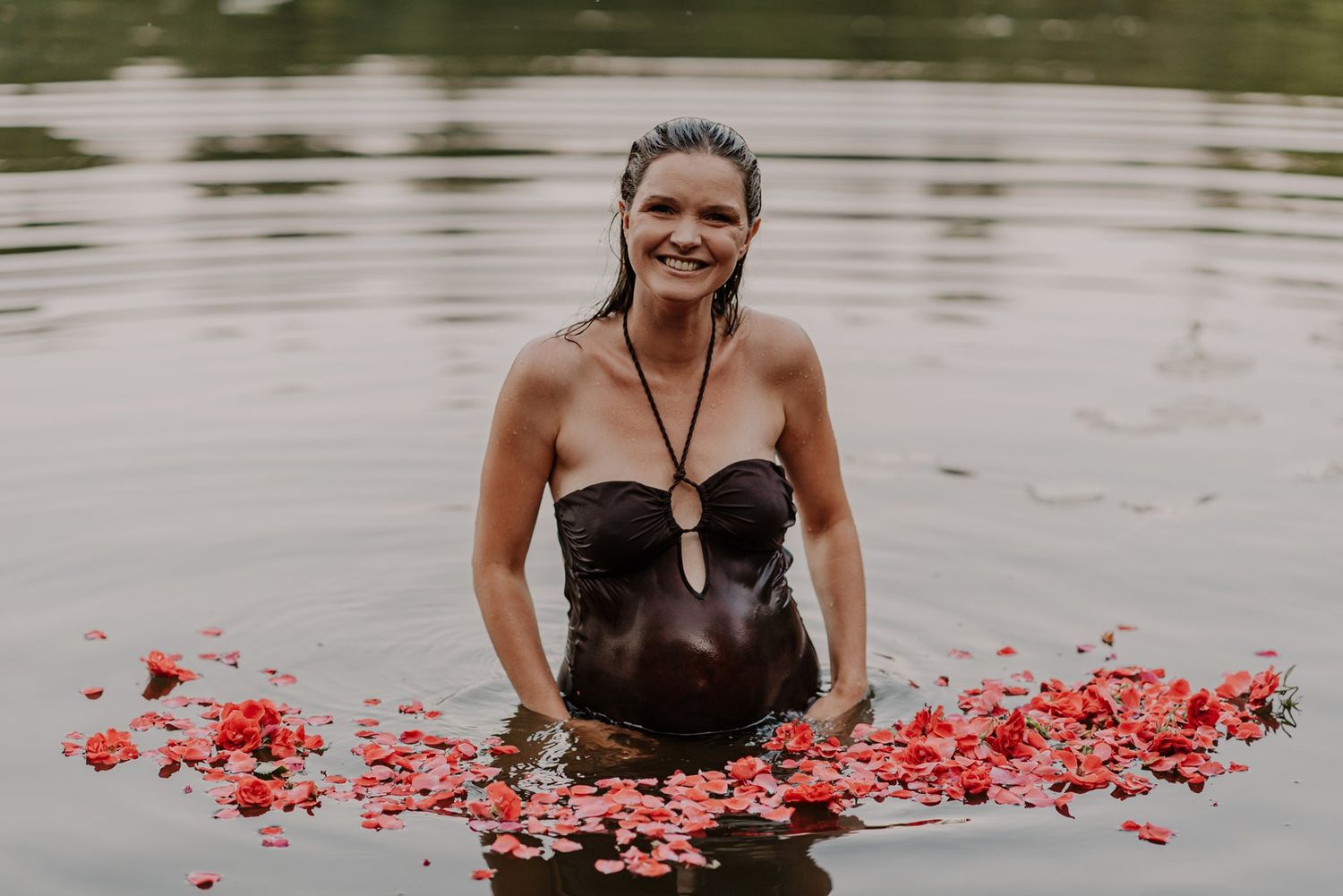 Person in brown dress poses in lake water surrounded by scattered red rose petals.