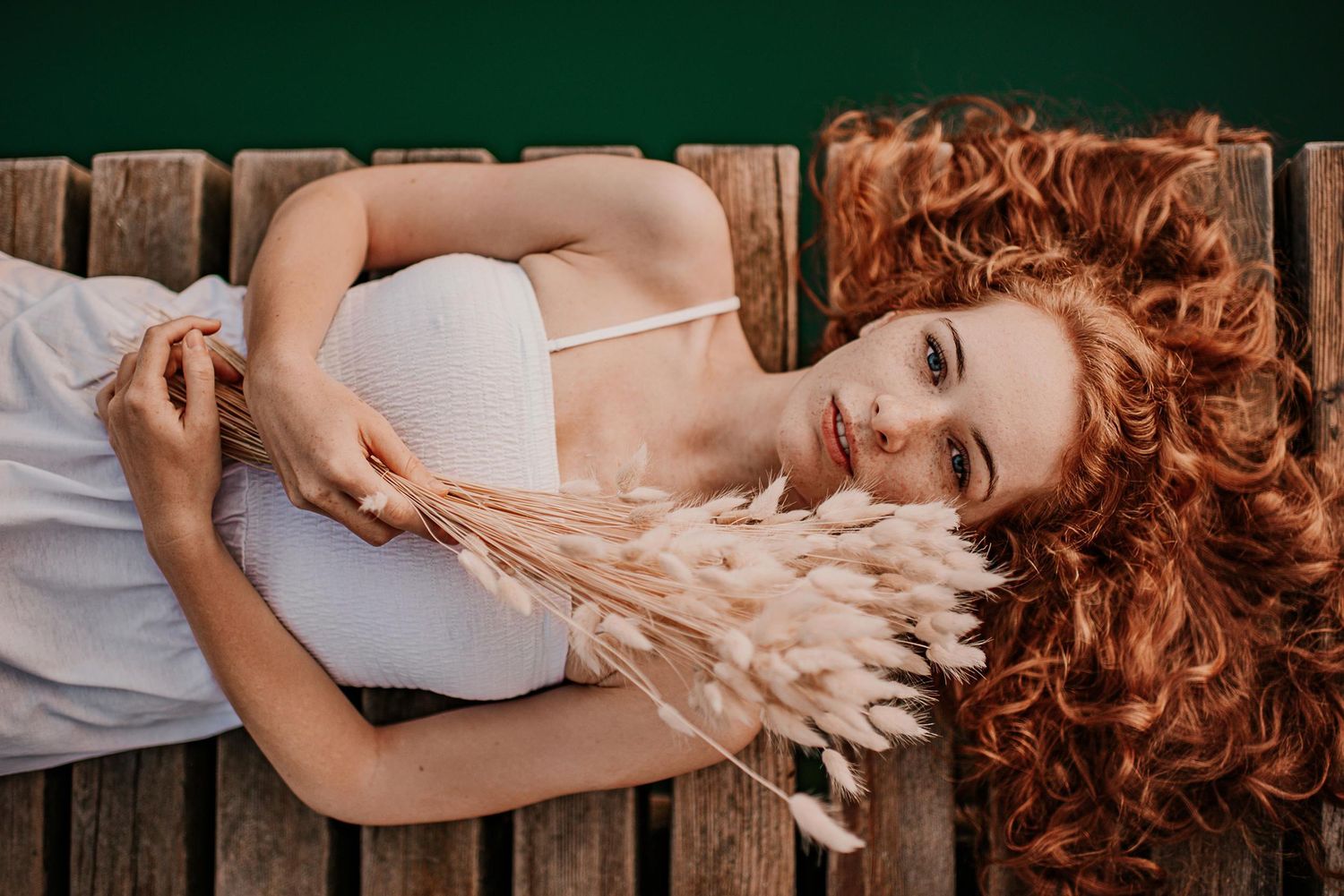 A person with red curly hair lying on a bed wearing a white dress in a romantic boudoir style photoshoot.