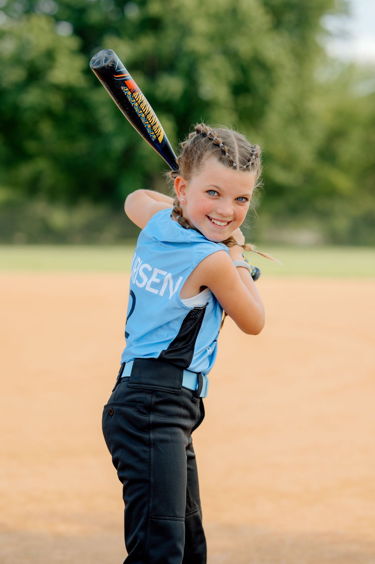 Young softball player in light blue jersey and black pants holds batting stance on sunny field.