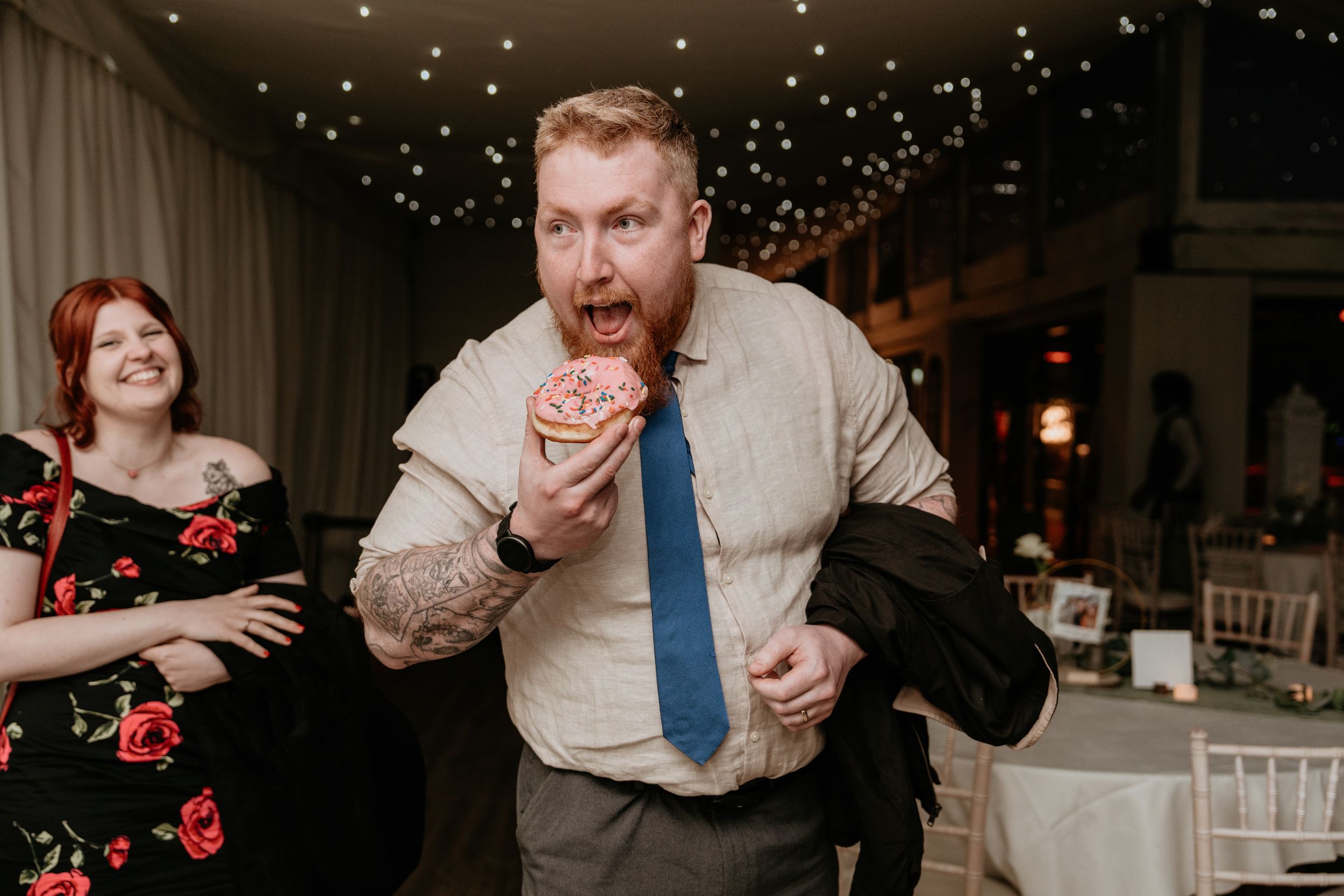 At a reception with twinkle lights overhead, a guest takes a big bite of food while dancing energetically.