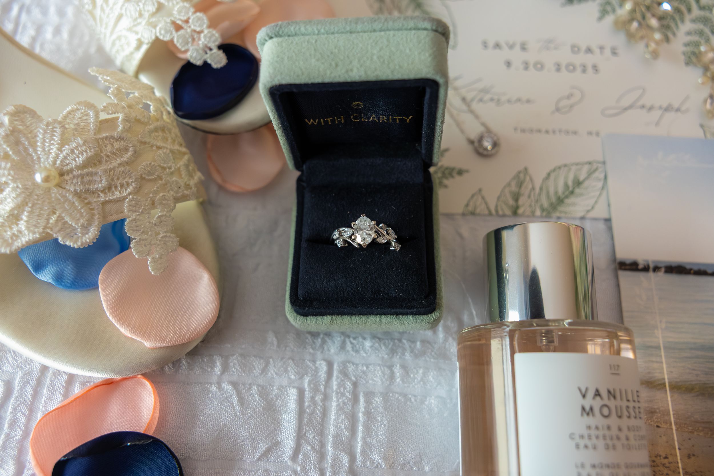 A diamond engagement ring in a dark velvet box displayed with ceramic heart trinkets and perfume bottle on white lace tablecloth.