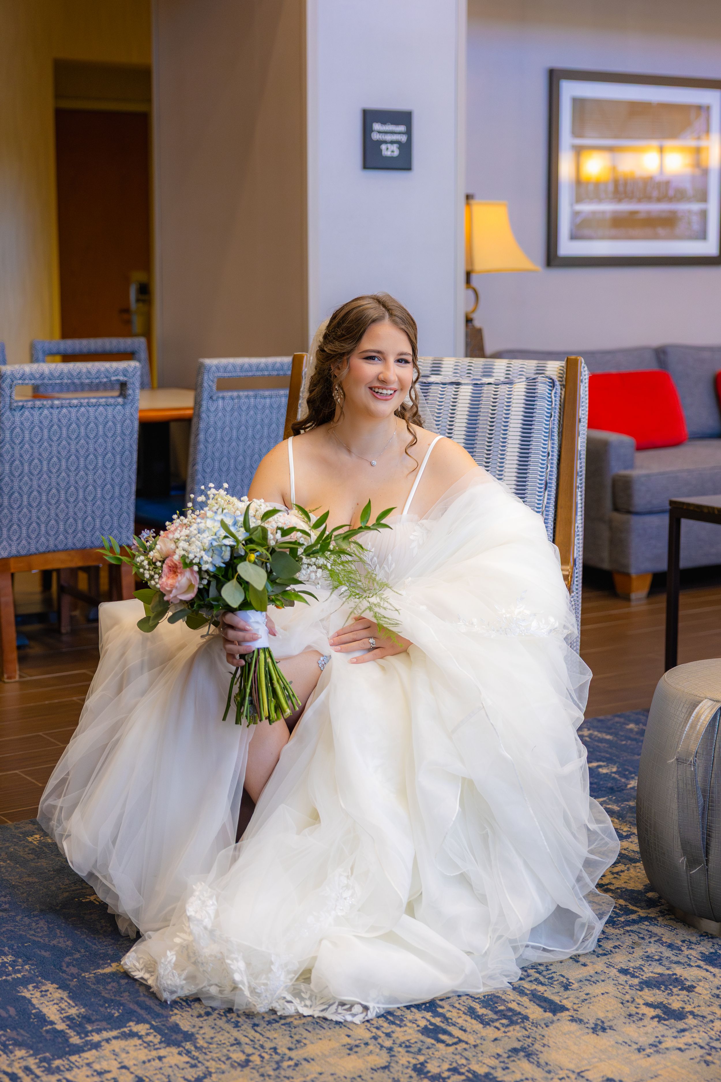 A bride in a flowing white wedding gown sits gracefully on a blue patterned carpet while holding a delicate floral bouquet.