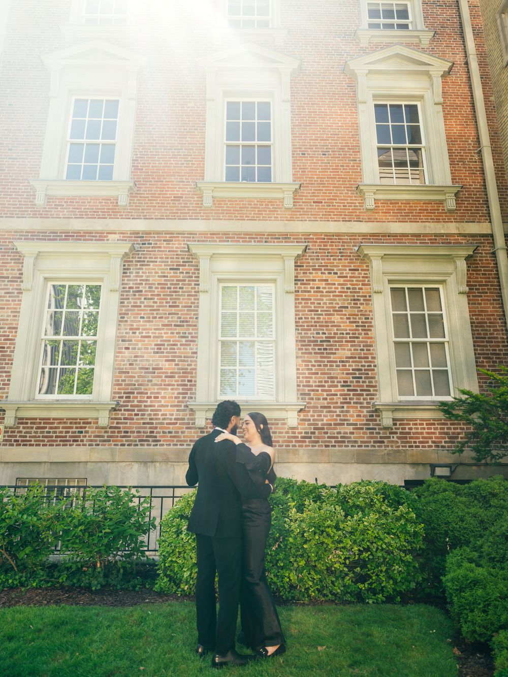 A couple embraces in front of a classic brick university building with white trim windows.