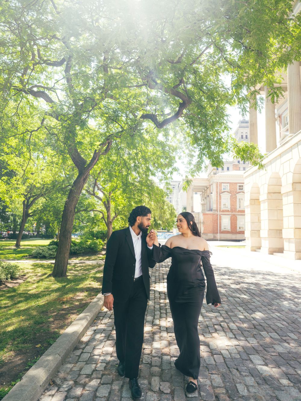 People in formal attire converse on an outdoor stone patio surrounded by green trees on a sunny day.