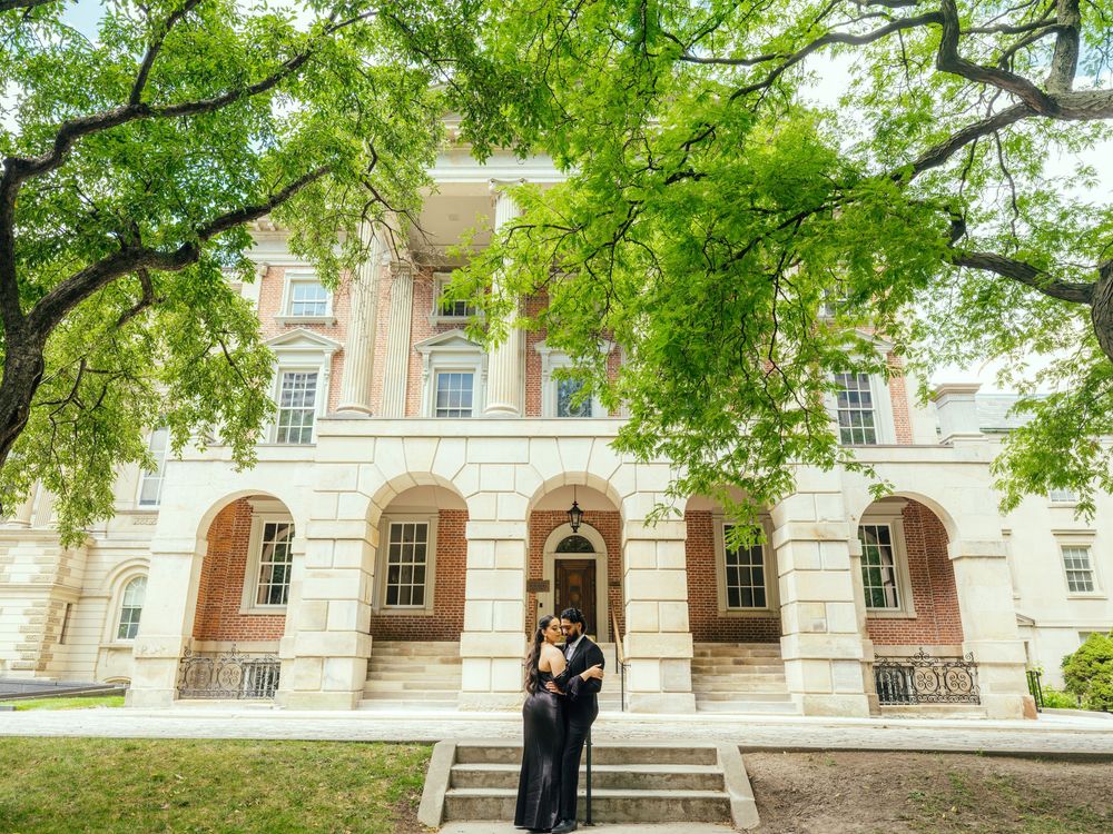 Elegant couple poses beneath green trees outside historic colonial building with white columns and grand architecture.