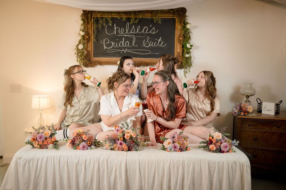 Five bridesmaids in satin robes celebrate at Chelsea's Bridal Suite party, holding bouquets of colorful flowers on a bed.