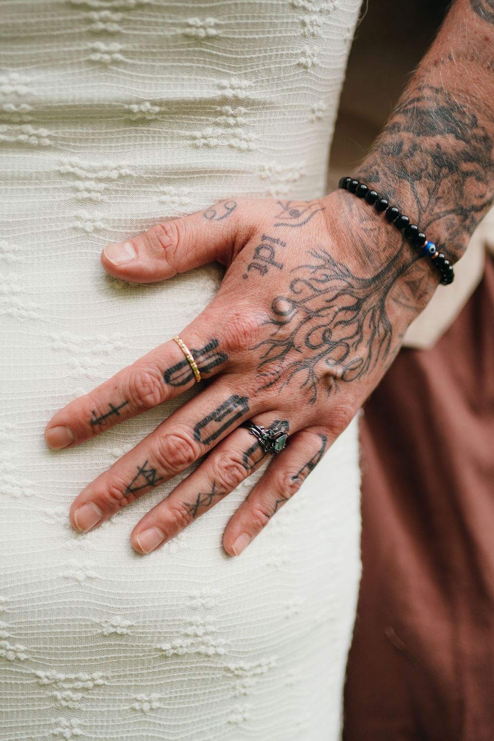 Close-up of a heavily tattooed hand wearing multiple rings and a black beaded bracelet resting on a white textured dress.