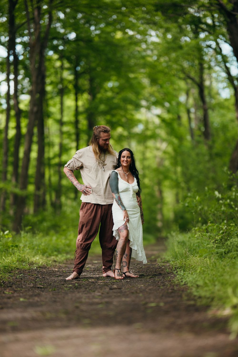 Couple in bohemian attire standing on a forest path, man in linen shirt and woman in white dress.