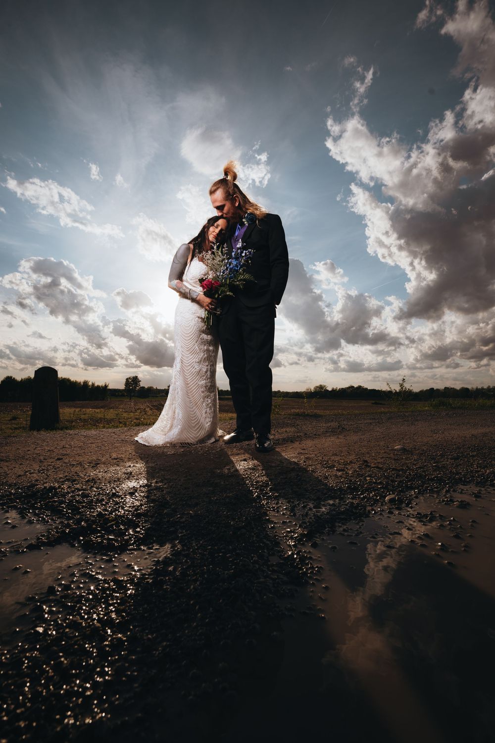 Bride and groom standing together under dramatic cloudy sky, bride holding colorful bouquet in outdoor wedding portrait.