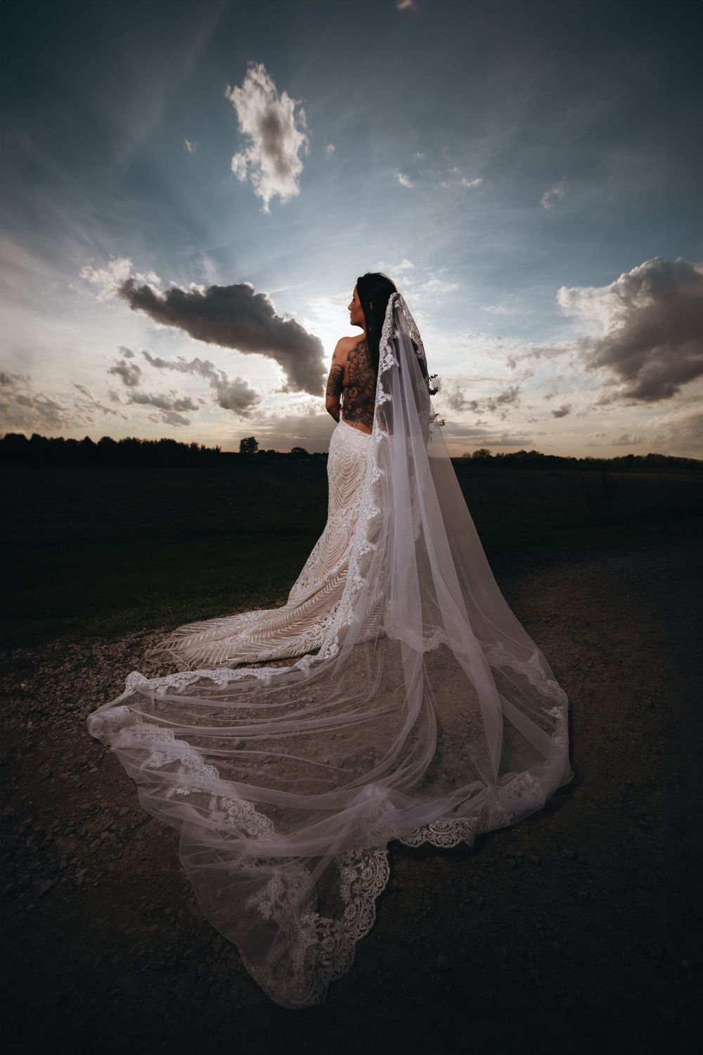 Bride in lace wedding gown and long veil stands against dramatic sunset sky on outdoor path.