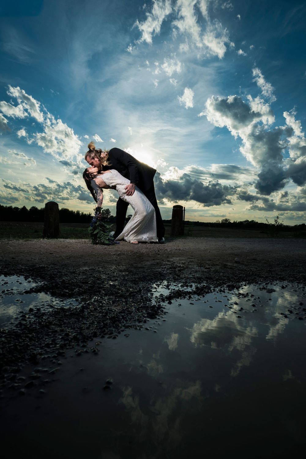 Bride and groom share romantic dip kiss at sunset with dramatic cloudy sky reflected in puddle below.