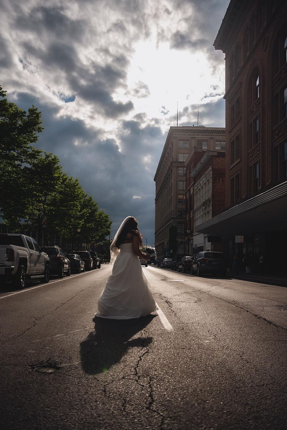 Dramatic silhouette of a bride in a wedding dress against moody urban architecture and cloudy sky.
