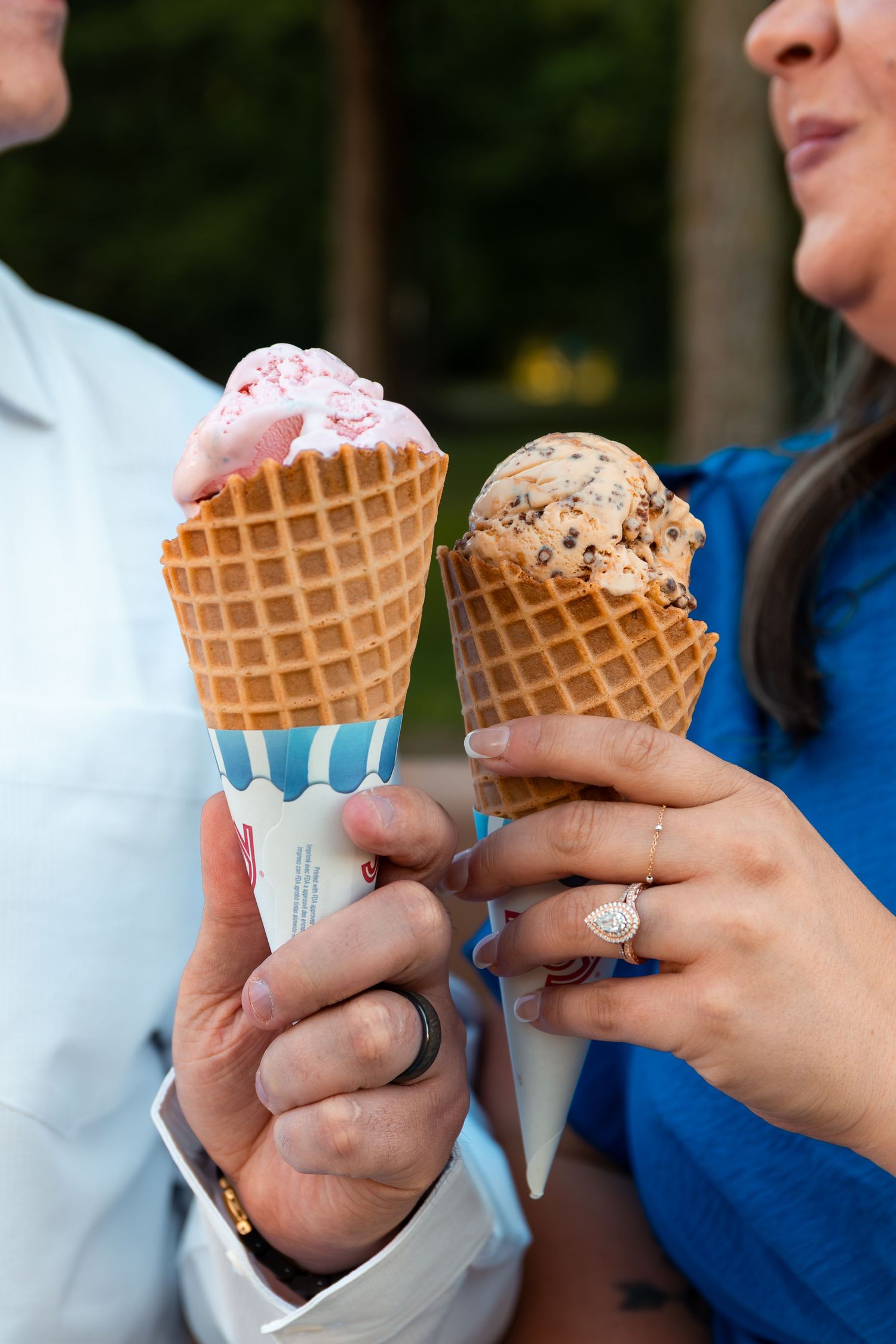 Couple holding waffle ice cream cones, woman wearing diamond engagement ring, enjoying strawberry and chocolate chip flavors.