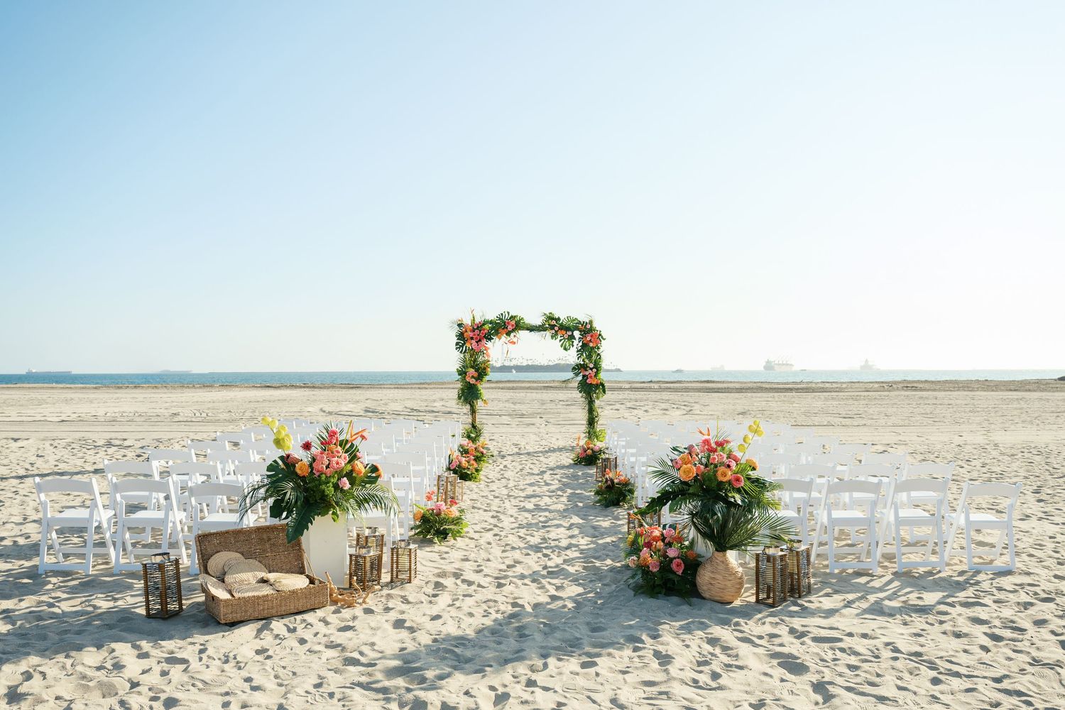 A romantic beach wedding setup with bamboo arch, flowers, and chairs arranged along white sandy shoreline.