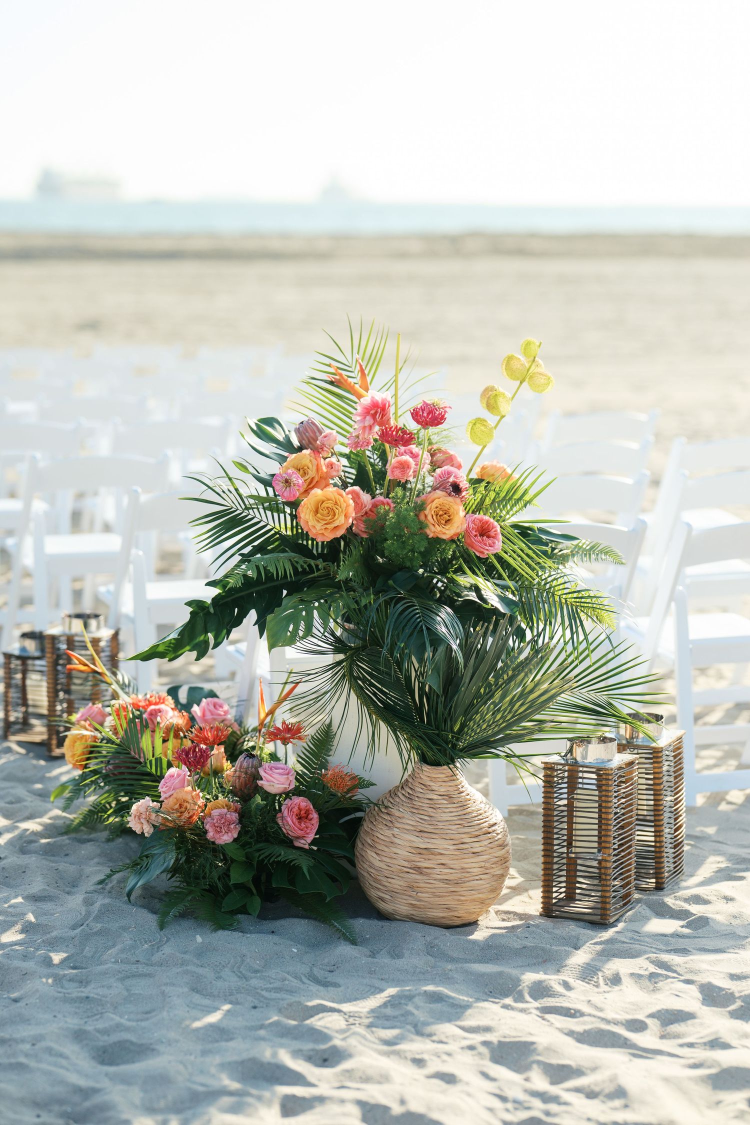 Tropical floral arrangements with coral roses and palm fronds decorate a sandy beach wedding setting with lantern accents.