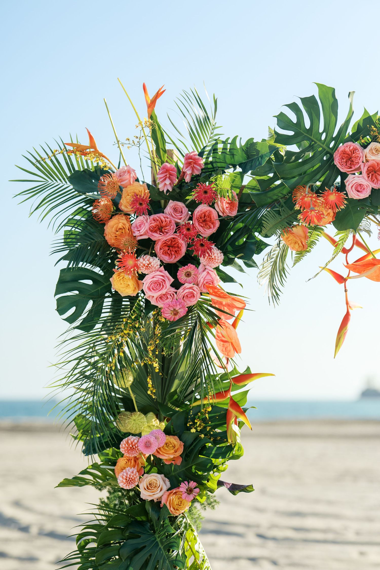 Tropical floral arrangement with pink roses, orange accents and monstera leaves displayed on a sandy beach.