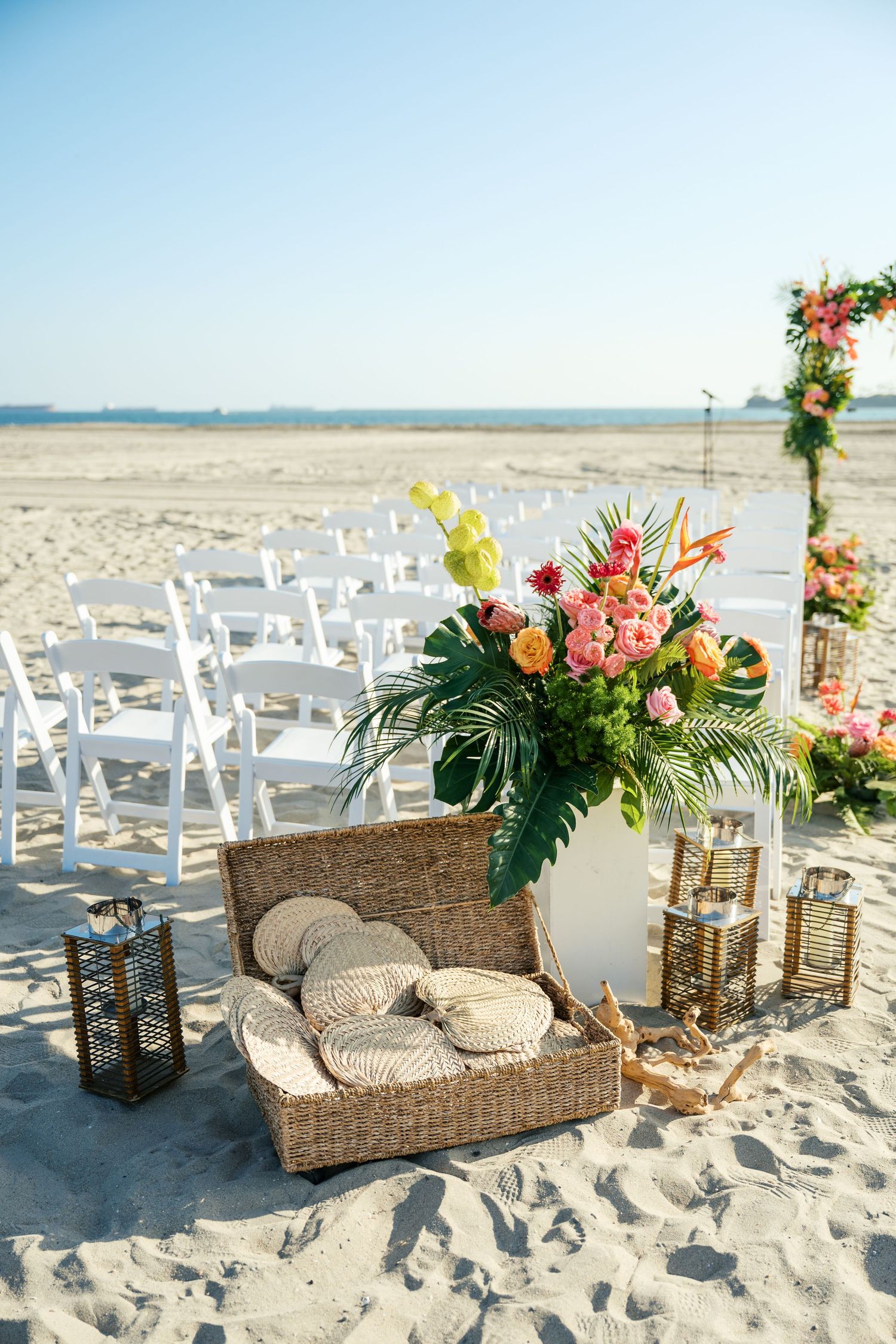 Elegant beach wedding setup with white chairs, woven seating, and tropical floral arrangements on sandy shores at sunset.