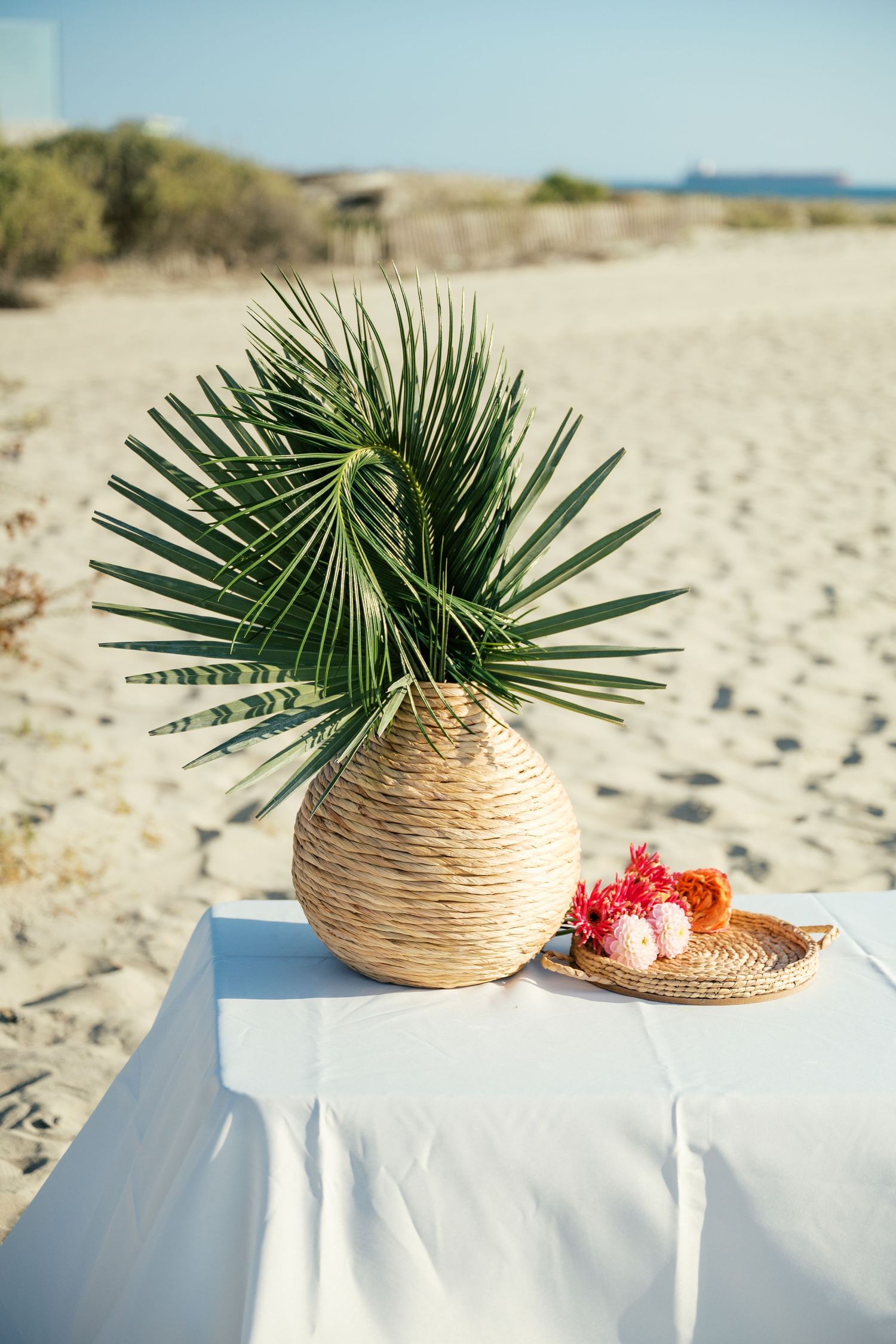 Tropical palm fronds displayed in woven straw vases against a beachy white sand background.