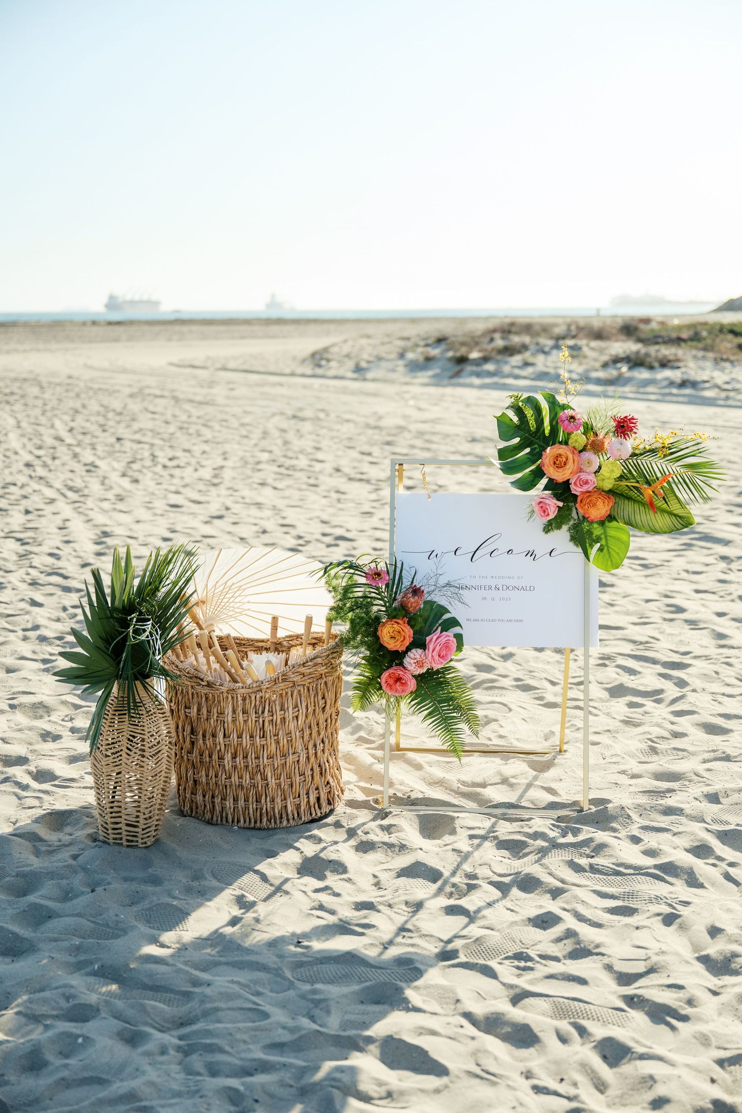 Tropical beach wedding decor featuring woven baskets, palm leaves, and pink flowers arranged on white sand.