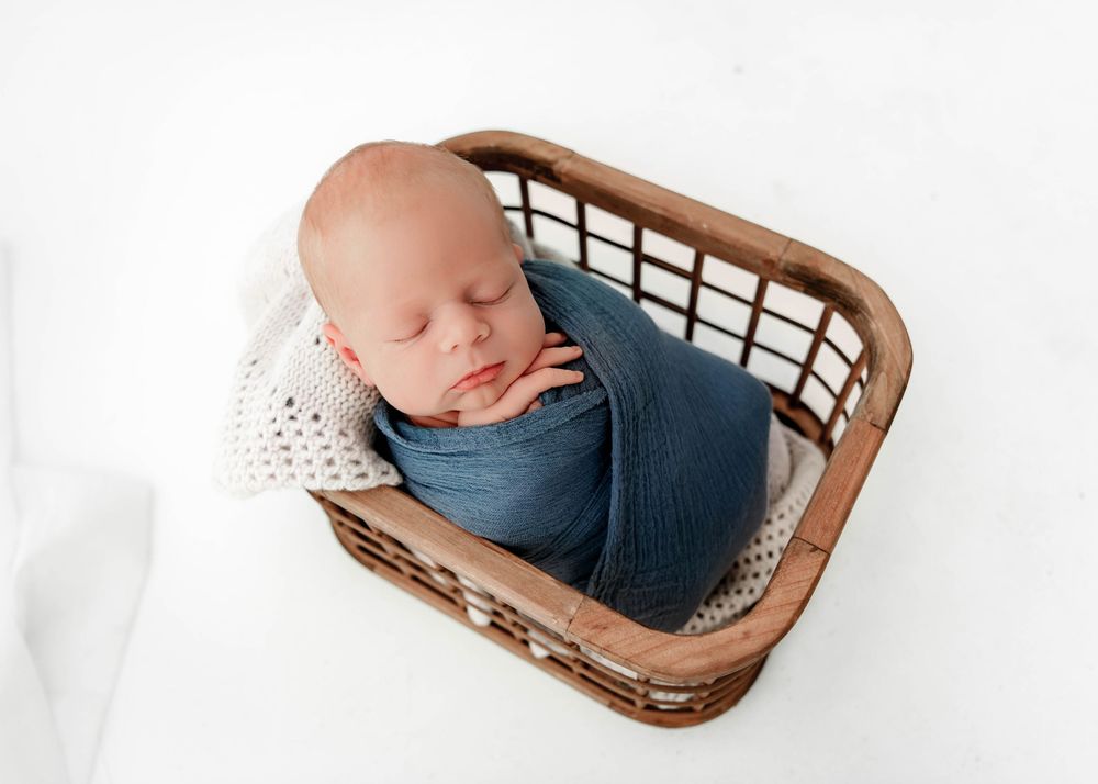 Newborn baby sleeping peacefully in woven basket with navy wrap and white knit blanket.
