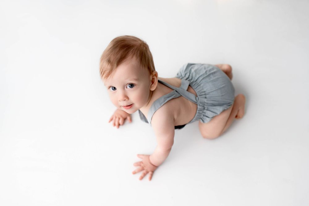 Infant in grey outfit crawling on white studio floor.
