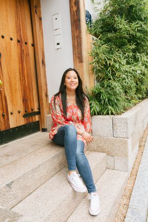 Person in casual outfit sits on stone steps near wooden door and bamboo plants.