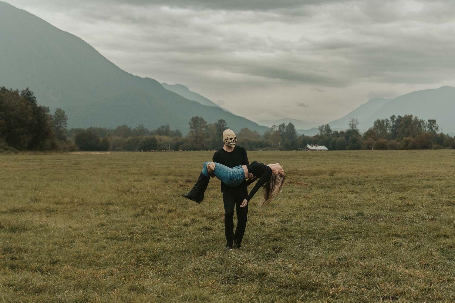 Spooky Halloween couple photoshoot with woman in black top, jeans and man in mask carrying her through foggy open field with mountains, WA