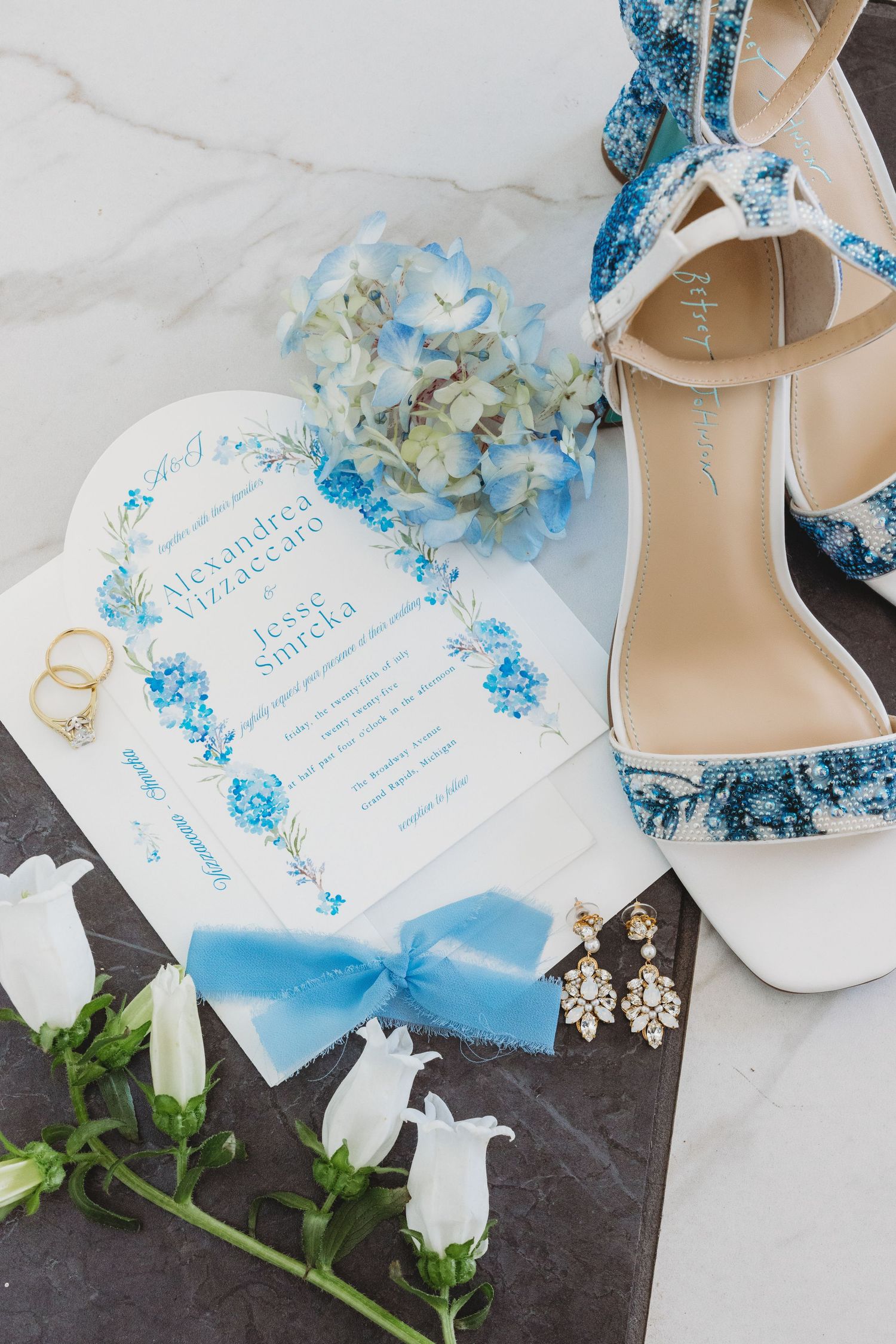 Wedding invitation suite with blue floral designs paired with elegant white heels and delicate flowers on dark surface.