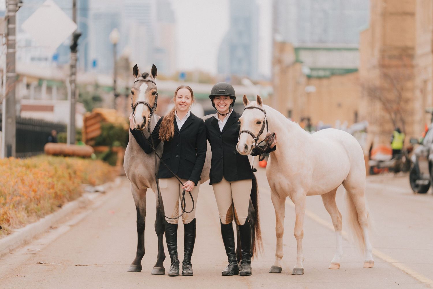 Two equestrians in riding gear stand with their horses on an urban street during autumn.