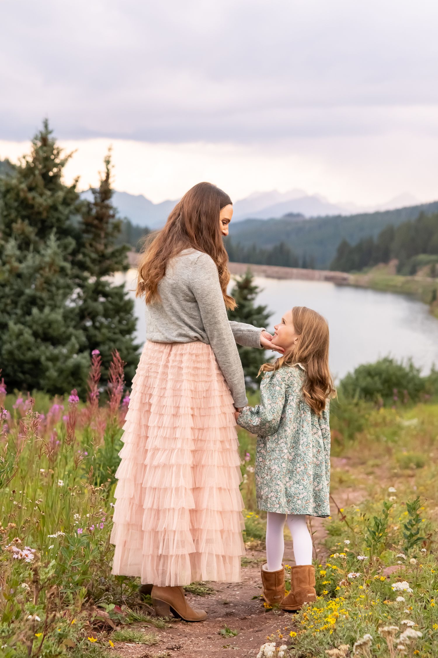 Parent and child sharing a sweet moment in a field of pink wildflowers by a mountain lake.