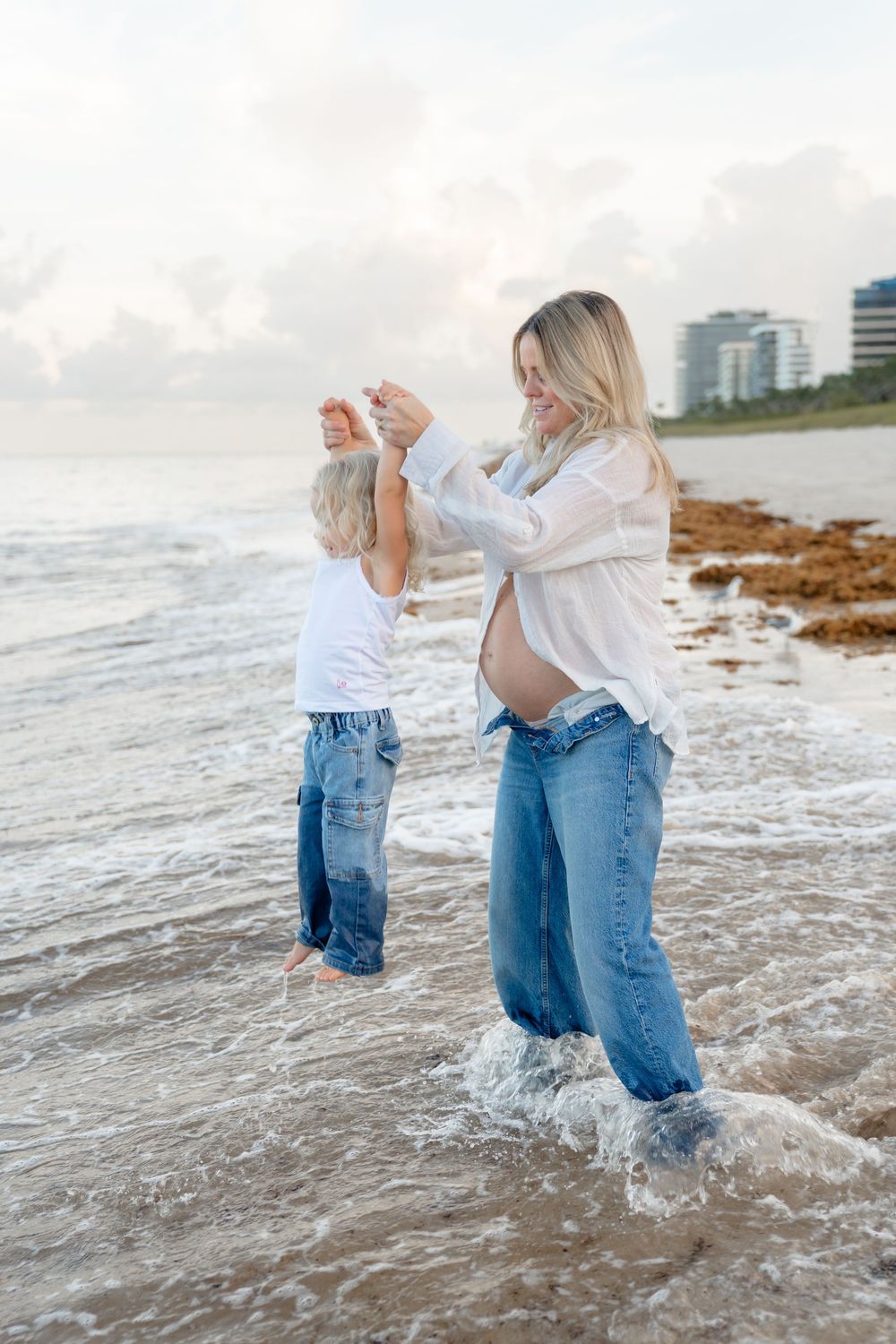 Two people in jeans and white tops play in the ocean waves at sunset.