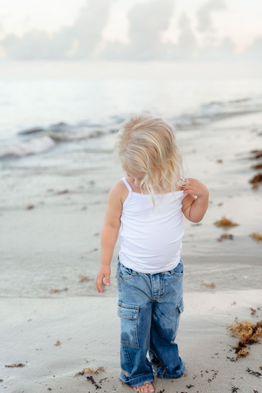A small child in jeans and white tank top exploring a sandy beach covered in seaweed.