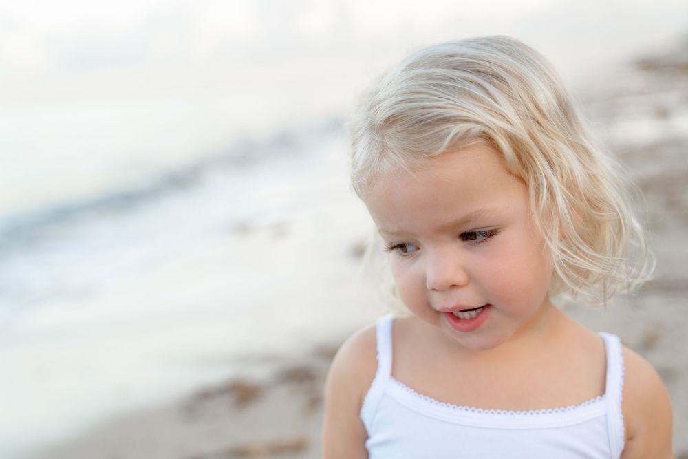 A young child with blonde hair wearing a white tank top smiles at the beach with blurred ocean background.