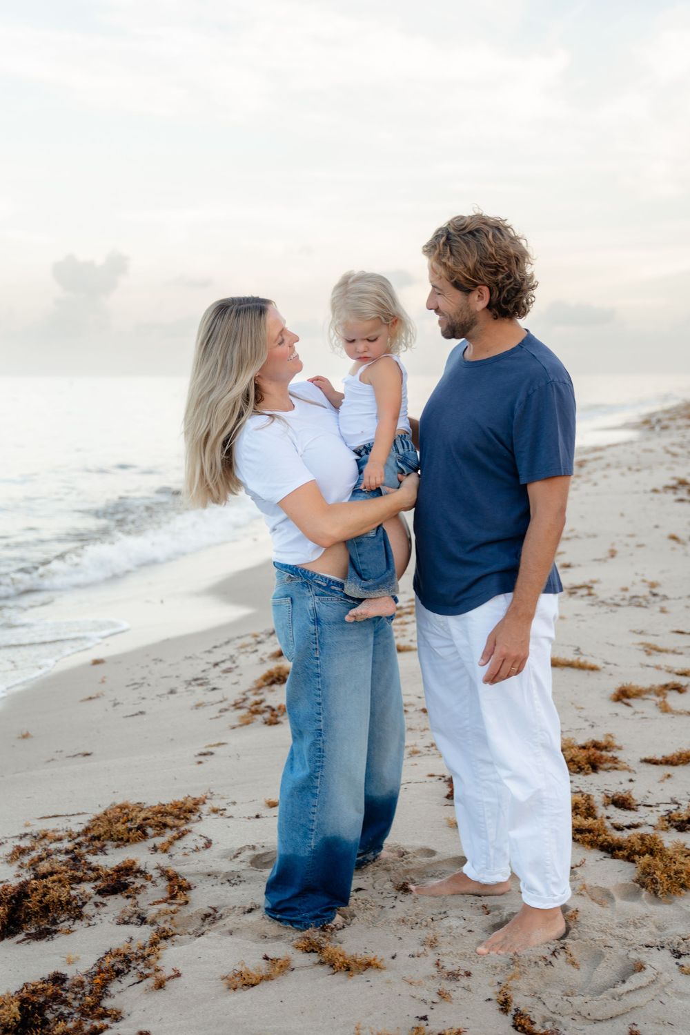 A family shares tender moments together on a beach with seaweed and ocean waves in the background.