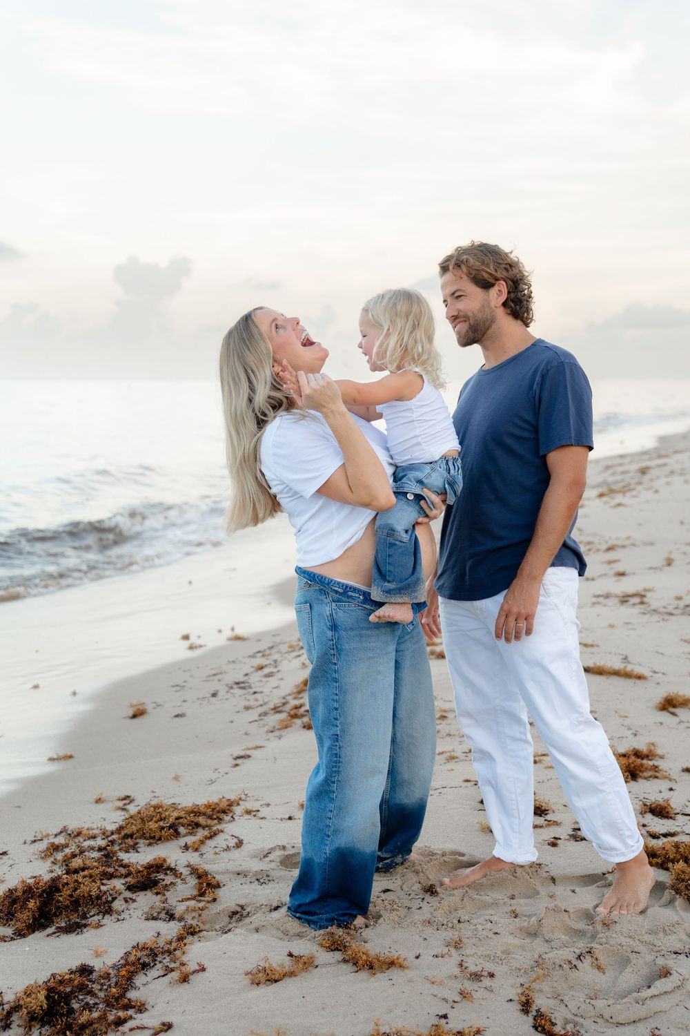 Family enjoying playful moments together on a sunny beach with waves and seaweed in the background.