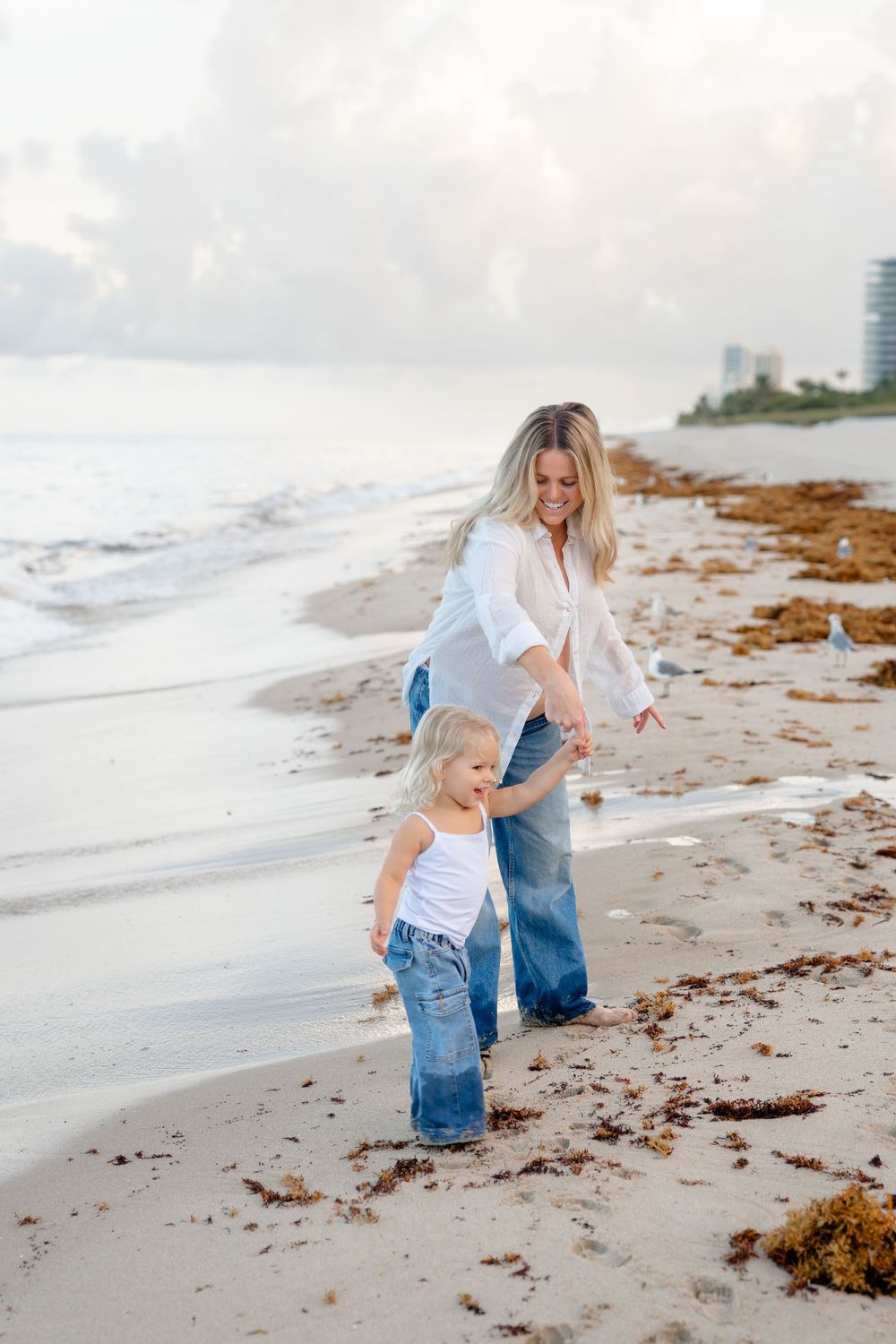 Two people in casual denim and white outfits share a joyful moment while walking along a beach at sunset.
