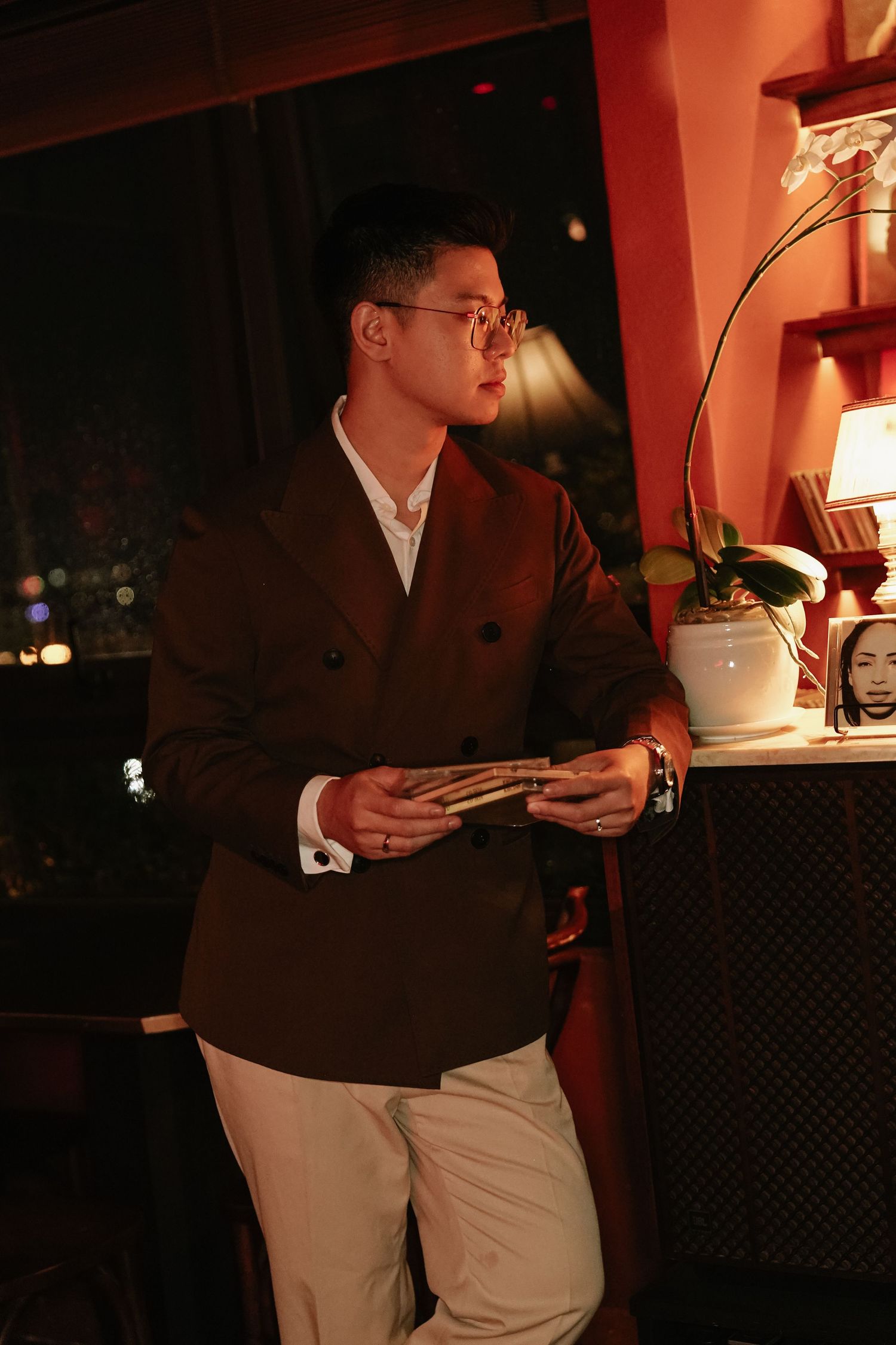 Stylish man in brown double-breasted blazer holding a book in a warmly lit evening setting.