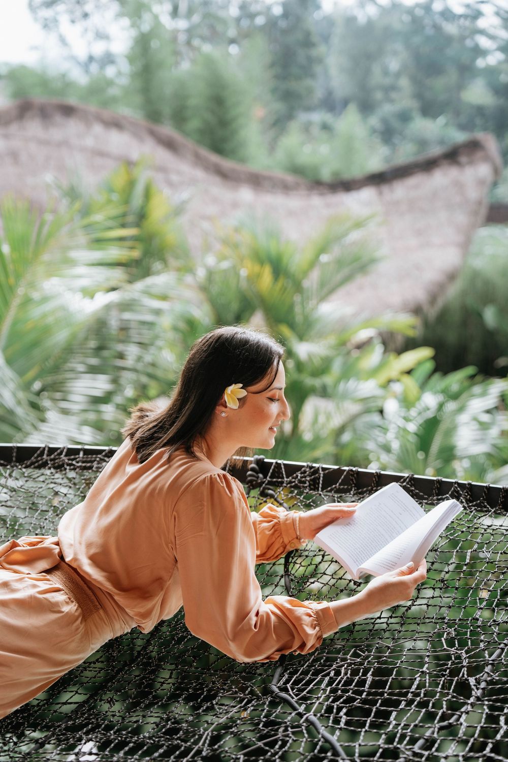 Person reading while lying on a net hammock in a tropical setting.