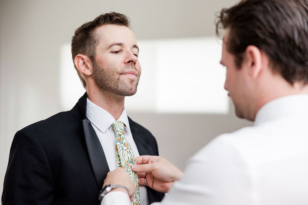 A groom getting ready for his wedding as someone helps adjust his tie and collar in a bright indoor setting.