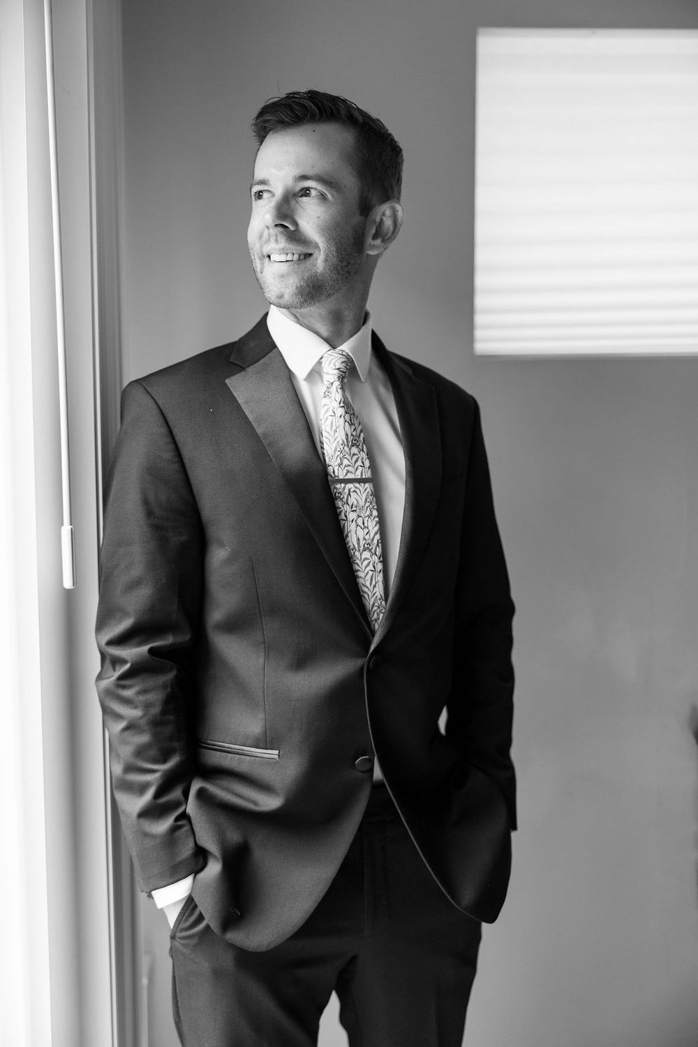 Professional black and white portrait showing a person in a dark suit looking out a window.